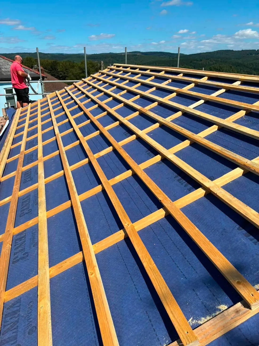 A roofer fitting battens and membrane to a large barn roof, with Forest of Dean hillside views behind