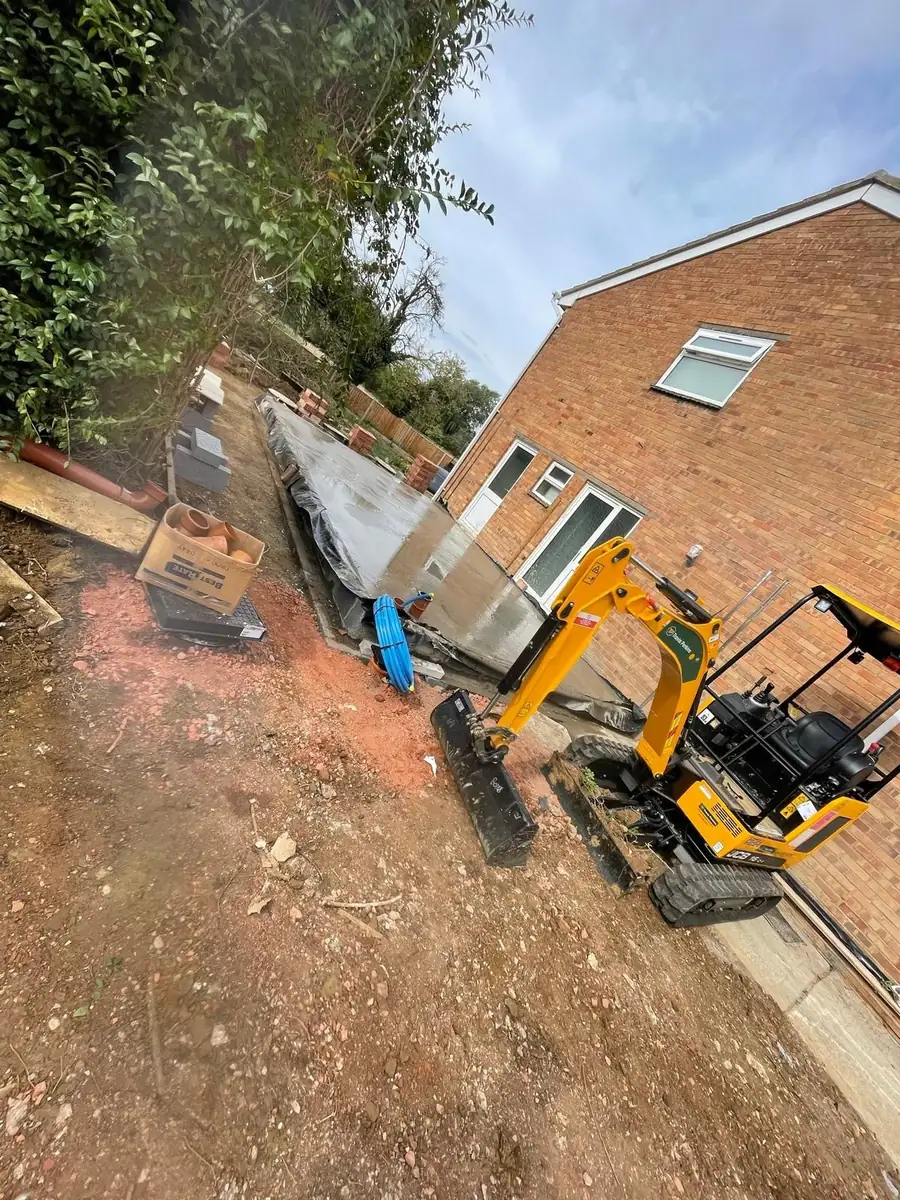 A mini digger manoeuvred through a side access route into a rear garden, with new footings visible alongside the house