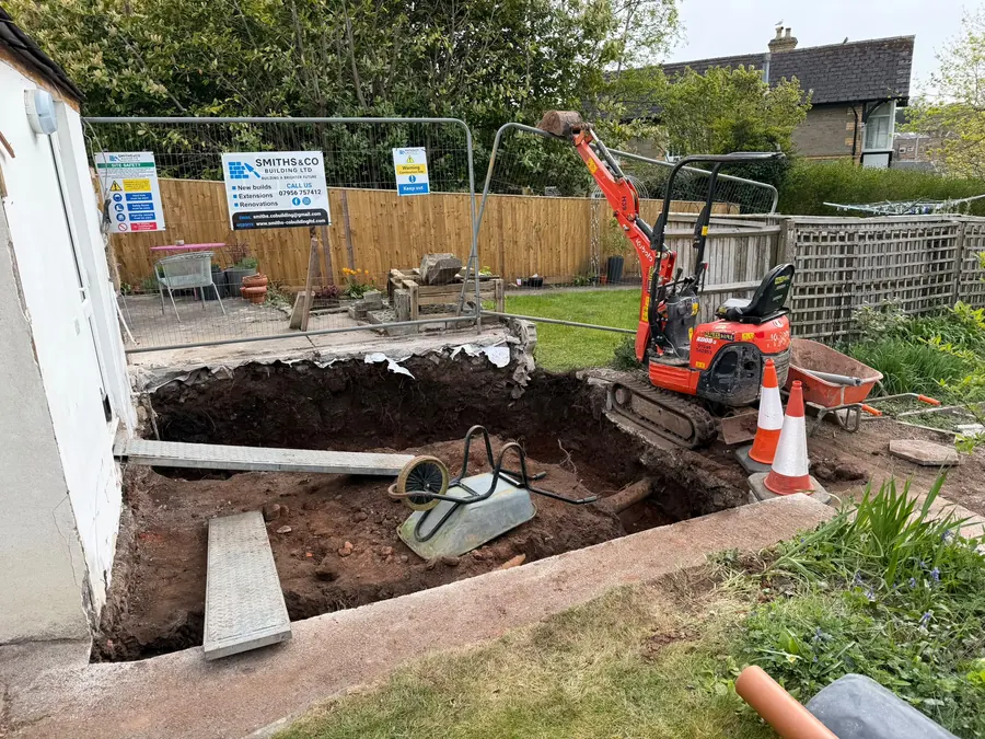 A dug-out site with a Smiths & Co site board on the fence, mini digger and spoil heaps alongside the trench