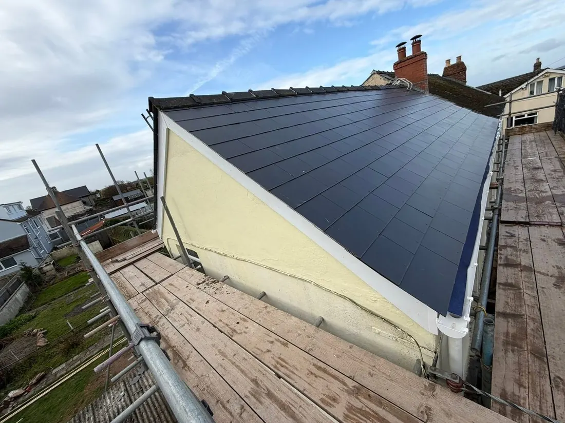 Newly tiled dark grey roof with a rendered gable end and chimney stacks behind, viewed from scaffolding